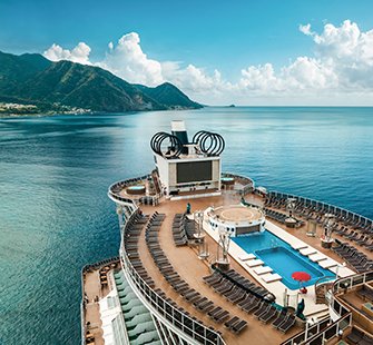 A cruise ship deck featuring a pool and sun loungers with mountains in the background | MSC Cruises A cruise ship deck featuring a pool and sun loungers with mountains in the background | MSC Cruises