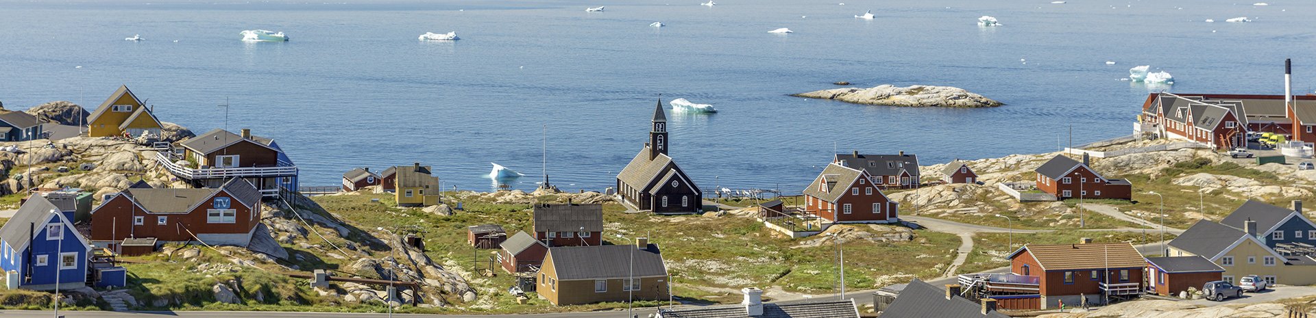 Colorful houses and a church overlooking an Arctic coastline with icebergs in the distance | MSC Cruises Colorful houses and a church overlooking an Arctic coastline with icebergs in the distance | MSC Cruises