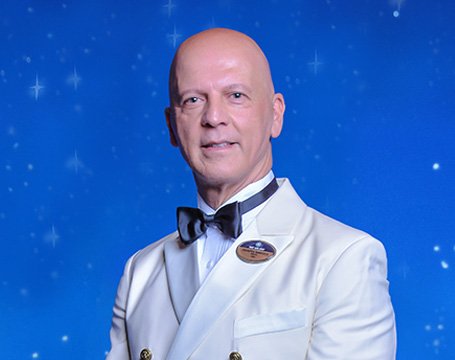 A crew member in a white formal uniform with an MSC Cruises badge against a starry blue background | MSC Cruises A crew member in a white formal uniform with an MSC Cruises badge against a starry blue background | MSC Cruises