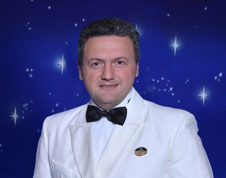A crew member in a white formal uniform with a bow tie against a starry backdrop | MSC Cruises A crew member in a white formal uniform with a bow tie against a starry backdrop | MSC Cruises