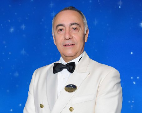 A crew member in an elegant white uniform with a name badge against a starry blue background | MSC Cruises A crew member in an elegant white uniform with a name badge against a starry blue background | MSC Cruises