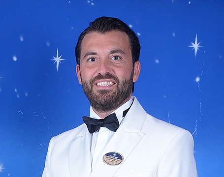 Crew member in formal white uniform with MSC badge against a starry blue background | MSC Cruises Crew member in formal white uniform with MSC badge against a starry blue background | MSC Cruises