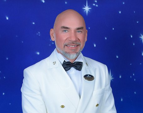 A crew member in a white uniform with a bow tie against a starry backdrop | MSC Cruises A crew member in a white uniform with a bow tie against a starry backdrop | MSC Cruises