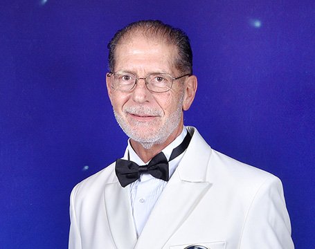 A man in a white tuxedo jacket with an MSC Cruises badge against a starry blue background | MSC Cruises A man in a white tuxedo jacket with an MSC Cruises badge against a starry blue background | MSC Cruises
