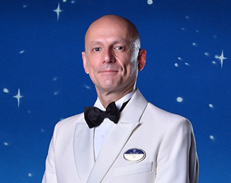 Crew member in a white uniform with a bow tie against a starry backdrop | MSC Cruises Crew member in a white uniform with a bow tie against a starry backdrop | MSC Cruises