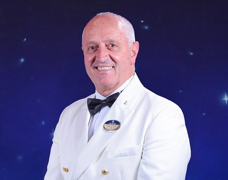 A crew member in formal white uniform with a name badge against a starry backdrop | MSC Cruises A crew member in formal white uniform with a name badge against a starry backdrop | MSC Cruises