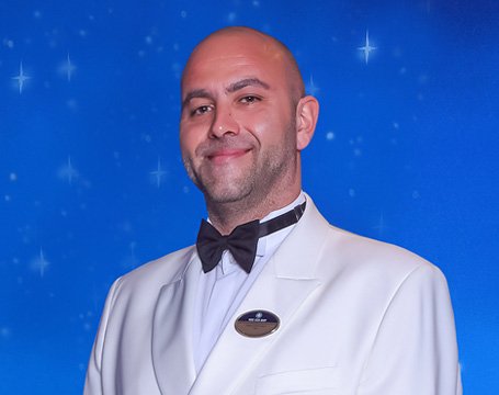 A crew member in a white uniform with an MSC name badge against a starry blue background | MSC Cruises A crew member in a white uniform with an MSC name badge against a starry blue background | MSC Cruises